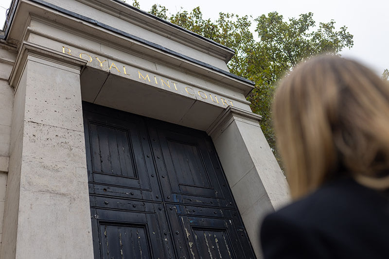 A presenter from the podcast strats up and the giant black wooden entrance doors to the Royal Mint site at Tower Hill