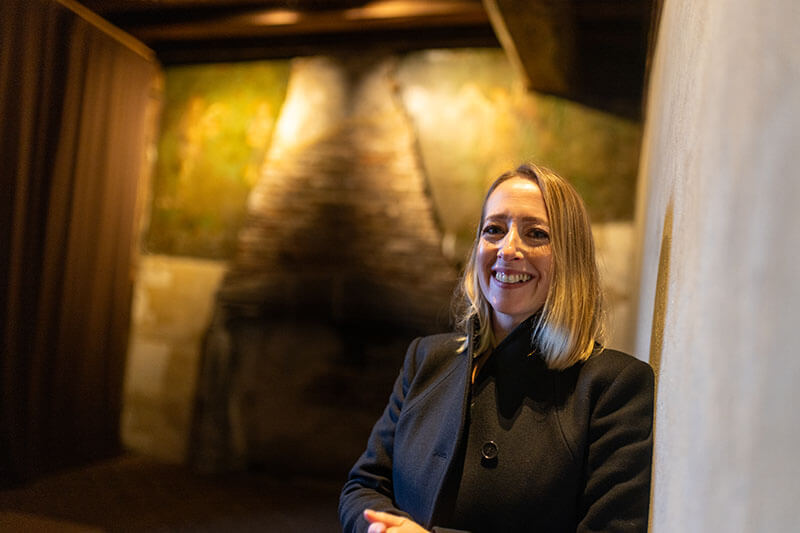 A female presenter with long blonde hair and a black coat stands smiling in front of a medieval wall painting