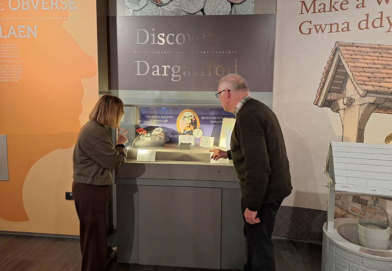 Photograph of Royal Mint Museum Exhibition manager Abigail Kenvyn interviewing Bob Meggs in front of an exhibition of his mother's maundy collection.