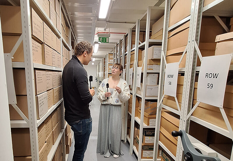Two people standing in a storage room full of shelves of cardboard boxes, each with a microphone in theirs hand