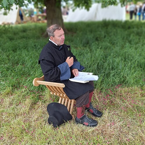 A picture of numismatist Jeremy Burrows seated on a wooden chair in a field at the re-enactment of the battle of Naesby. He is wearing black civil war era clothing.
