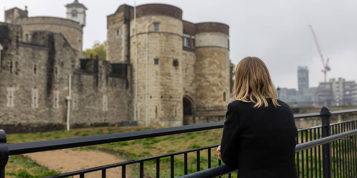 Blond haired woman looking over a railing over what used to be a moat but is now grass. In the background are the huge stone buildings that make up the Tower of London