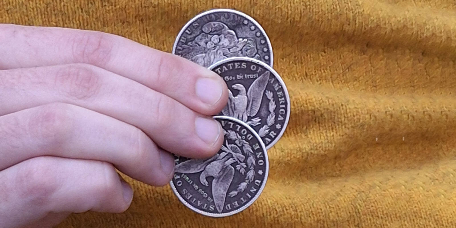 A hand holding three large silver coins fanned out as if to do a magic trick. They have eagles on the side that is showing and the words United States of America around the outside.