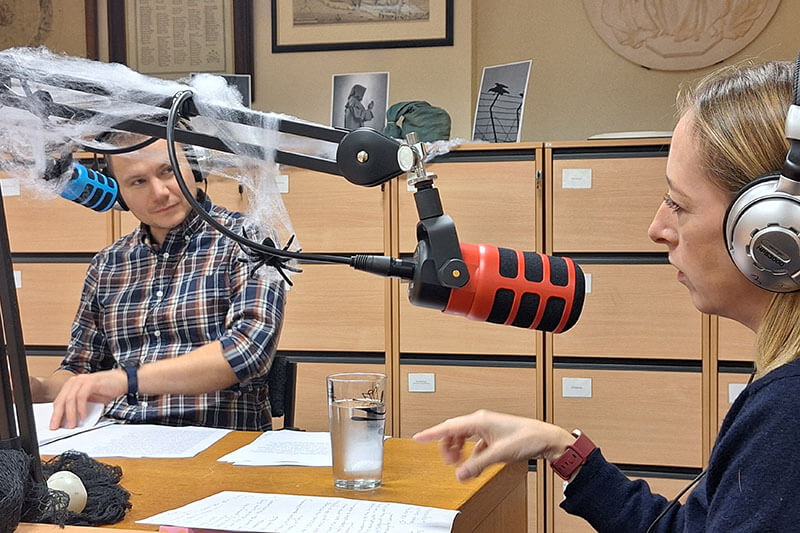A man and a woman with headphones on speak into microphones while the studio is covered in pretend spiders, skulls and spider web